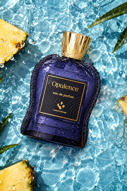 A bottle of Opulence Eau de Parfum by Luxury Couture, featuring a deep blue-purple glass body with a metallic gold cap, set against a background of rippling blue water with pineapple slices and green leaves.