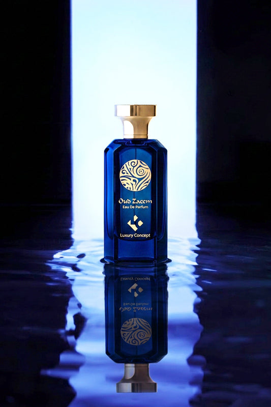 A luxury blue glass perfume bottle with a gold cap and gold logo, standing on a reflective dark surface with a bright vertical light behind it.